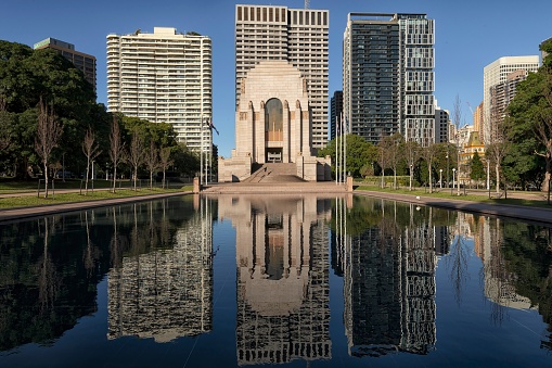 Sydney, New South Wales, Australia: Northern side of Anzac Memorial in Hyde Park with pool of reflection reflecting the memorial and Sydney city high rise buildings. Deep blue sky.