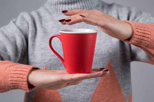 In female hands there is a red cup with a hot drink, one hand is above the cup, the concept of warmth in the cold season, selective focus