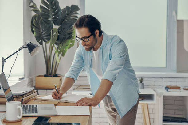 istock Confident young man making some notes while standing near his working place in office 1424878308