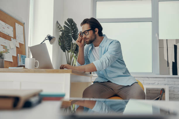 istock Confident young man using laptop while sitting at his working place in the office 1424880659