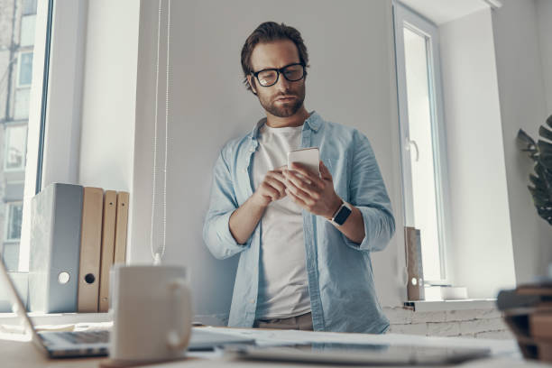 istock Confident young man using smart phone while standing near his working place in office 1424882693