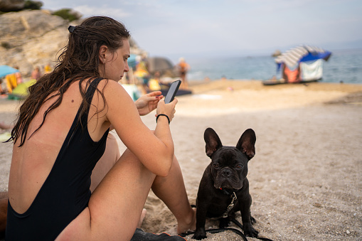 French Bulldog sitting next to young woman at beach while she's using smartphone