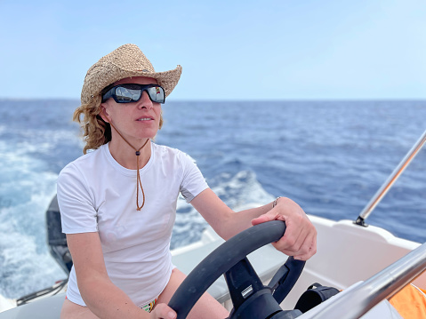 a smiling woman with sunglasses and straw hat, driving a sports boat on a blue water sea on a summer vacation day, selective focus, copy space