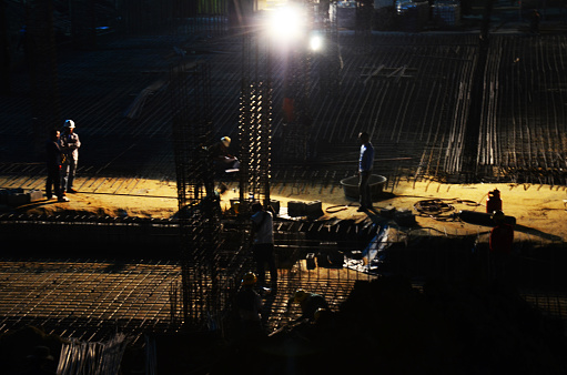 Asian labor people and thai labour worker knitting metal rods bars into framework reinforcement for concrete pouring for working structure building at construction site night time in Bangkok, Thailand