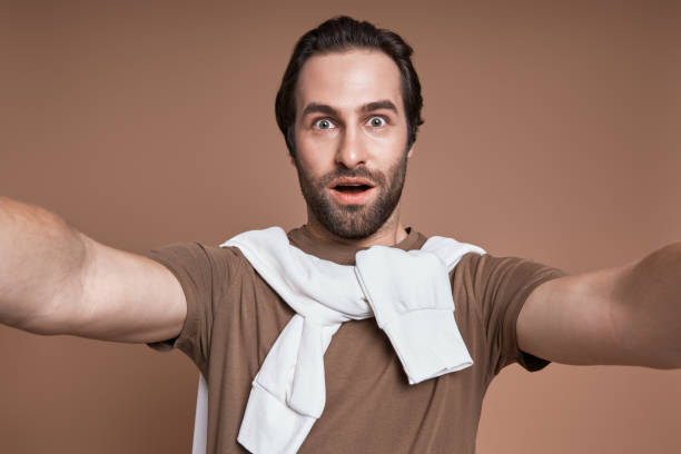 istock Surprised young man making selfie while standing against brown background 1424896864