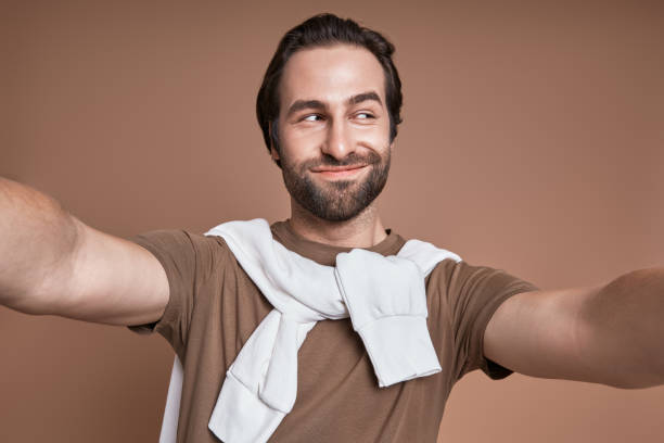 istock Playful young man making selfie and grimacing while standing against brown background 1424896910