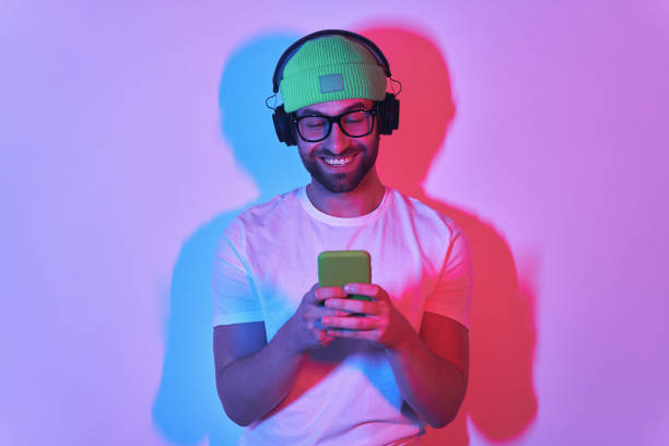 istock Handsome young man in headphones using smart phone and smiling against colorful background 1424900511