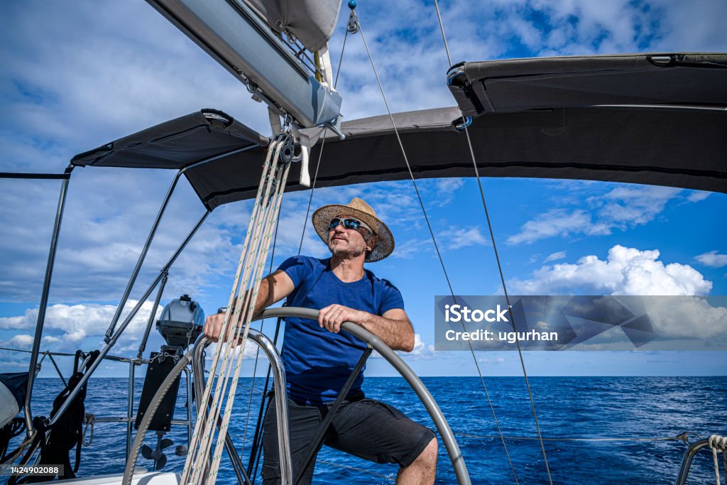 A man wearing a straw hat  is driving a sailboat and enjoying it. Sailing Stock Photo