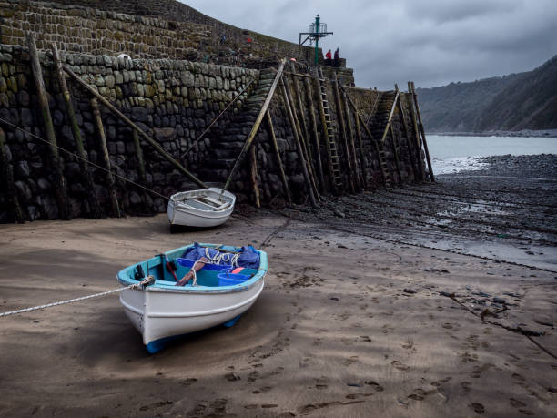 The harbour wall at Clovelly, Devon, during low tide. A white boat with a pale blue interior waits for the tide. Two people brave the cold to watch the sea, one wrapped up warm against the winter weather in a red jacket. The beach is covered with lines of rusted chains, waiting for the fishermen’s boats to return with their catch.