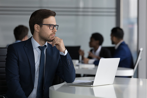 Thoughtful millennial businessman in glasses working at laptop computer in open office space, thinking over startup project ideas, creative decision, touching chin. Corporate meeting in background
