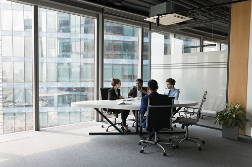 Diverse millennial business team talking in meeting room, negotiating on project at table at glass wall panoramic window, discussing deal in open space, modern office interior. Wide shot