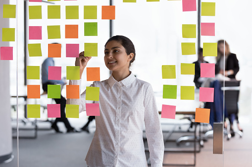 Happy confident young Indian business professional making project schedule, writing on paper sticky notes, working on strategy, business process at glass board in office