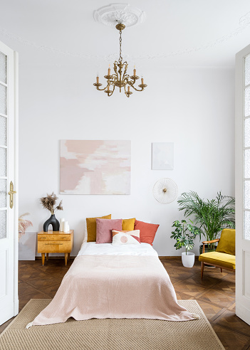 The style of the 60s concept. Bright vintage bedroom in apartment with retro interior design. Houseplants in room with authentic chandelier on a high ceiling over tidy bed, side table, armchair