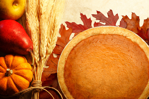 Autumn fruit and wheat stalks bound together by twine rest nest to a Thanksgiving day pumpkin pie.