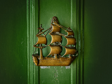 A vintage brass door knocker in the shape of a three-masted ship. The knocker rests against a green, wooden door.