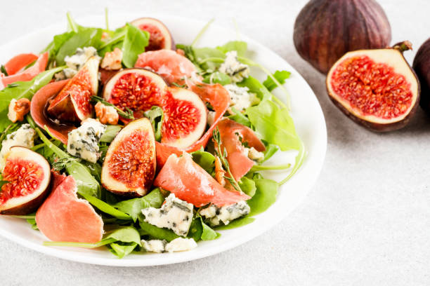 Healthy food - salad with fresh fig, blue cheese, prosciutto, arugula and spinach on white table background with ripe figs around. Selective focus Healthy food - salad with fresh fig, blue cheese, prosciutto, arugula and spinach on white table background with ripe figs around. Selective focus plate fig blue cheese cheese stock pictures, royalty-free photos & images