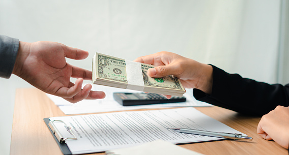 A bank employee offers dollars in a real estate investment loan with an approved contract application for mortgage and home insurance offerings. Borrowing for future investments and building credit.