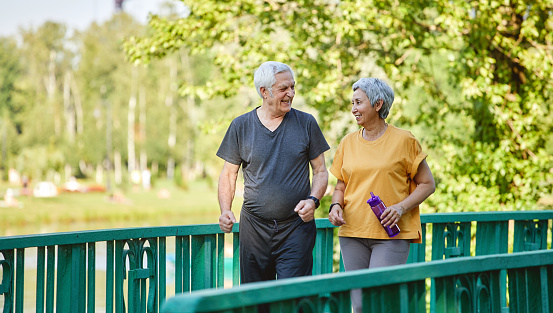 Asian and Caucasian middle-aged wife and husband wear activewear strolling along bridge in summer park, enjoy morning sportive walk together outside. Healthy lifestyle and retirement