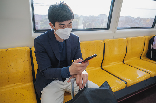 Asian businessman passenger man in suit wearing protective face mask using mobile phone and sitting inside BTS sky train or MRT underground train at railway platform while traveling to work, Bangkok, Thailand, Transportation and lifestyle concept