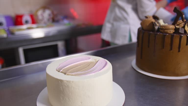 Pastry cook applies white cream with a spoon on top of the white cake. Close up. Chocolate cake at the table at backdrop.