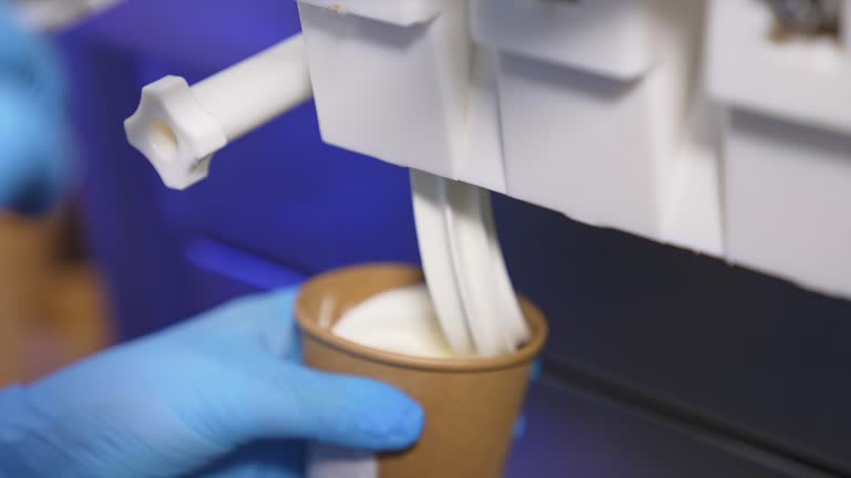 Hand in blue gloves puts a paper cup under a food automat. Soft creamy ice-cream is filling the cup close up.