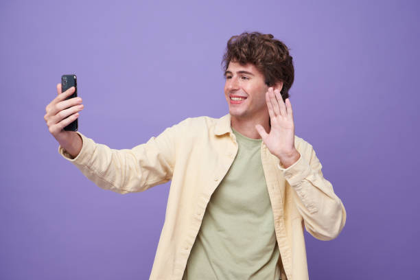 istock Young smiling handsome man saying hi and waving hand while making video call on smarphone isolated on purple banner background. 1424963059