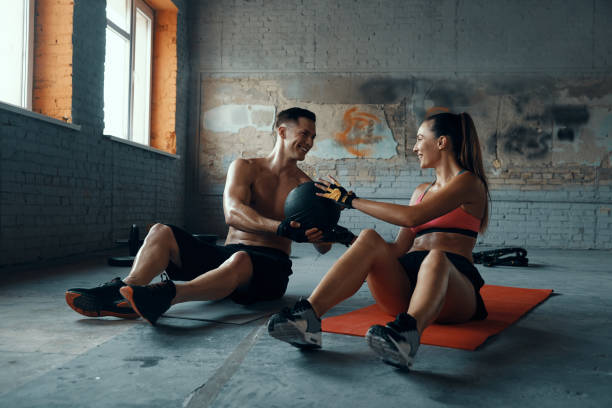 Happy fit couple exercising with medicine ball while sitting on exercise mats in gym stock photo