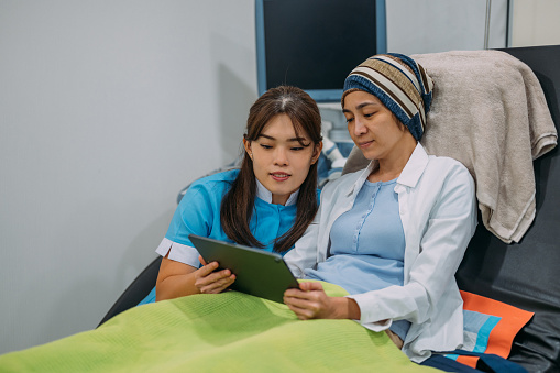 An Asian woman with cancer reviews test results with health worker. An Asian female gynecologist  showing cancer reviews and test results with her patient with digital tablet at hospital.