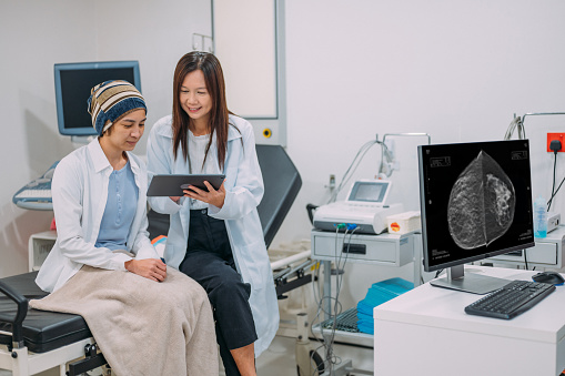 A Asian woman with breast cancer is meeting with her doctor. The doctor is showing her test results on an electronic wireless tablet. The doctor smiles as he gives good news regarding the patient's treatment.