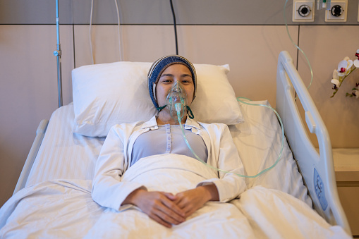 An Asian female cancer patient with oxygen mask lying on hospital bed. Sad female patient feeling sick at hospital. Depressed woman hospitalized in a medical clinic. Looking at camera. Cancer Patient Receiving Treatment and Psychological help concept