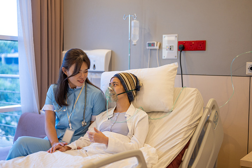 An Asian Chinese woman wearing a headscarf and oxygen mask with breast cancer patient is at a hospital. Patient feeling comforted by her female health care worker.