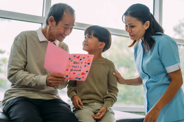 An Asian senior man received a Get Well card from his grandson sitting beside him at hospital ward. Senior male patient reading a Get Well card with his grandson sitting beside him at hospital ward. hospital card stock pictures, royalty-free photos & images