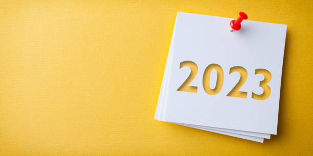 White Sticky Note With Happy New Year 2023 And Red Push Pin On Yellow Cardboard Background stock photo