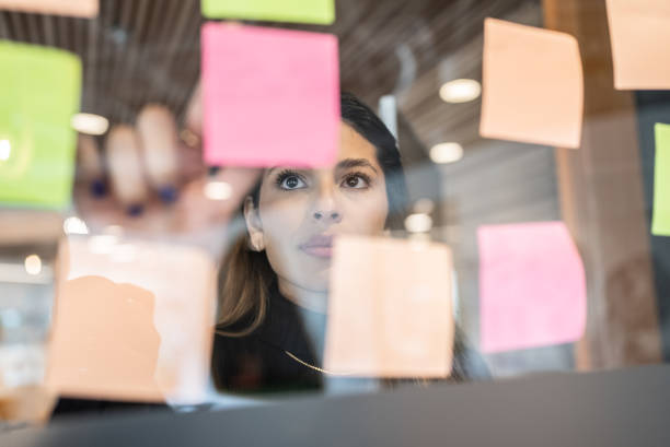 mujer joven escribiendo en post it en la oficina - setting goals fotografías e imágenes de stock