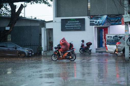 Adult thai in red raincoat is driving on motorcyce in hard rain in Bangkok Nawamin. Scene is in early evening in rainy season. In background are buildings and parked cars
