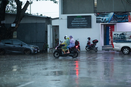 Two people in raincoats are driving on motorcyce in hard rain in Bangkok Nawamin. Scene is in early evening in rainy season. In background are buildings and parked cars