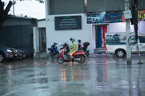 Thai person in raincoat is  driving on motorcyce  in hard rain in Bangkok Nawamin. Scene is in early evening in rainy season. In background are buildings and parked cars