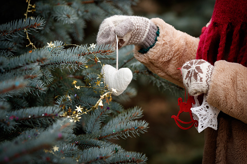 The hands of the girl in mittens are holding a New Year tree decoration. Decorated Christmas fir tree in the background. New Year and Christmas concept