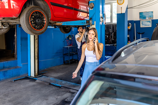 Young Car Mechanic is Repairing the Lifted Car while his Female Customer is Talking Using a Mobile Phone.