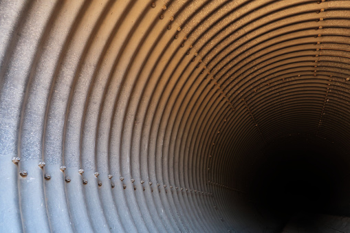 Close up of a large diameter corrugated galvanized steel culvert, viewing the inside at an angle, looking down along its side until it disappears into darkness in the distance.  Successive metal corrugated arcs/curves begin at left of horizontal frame and diminish into the center and top of frame until darkness overtakes them at lower right of frame.  A row of rivets is distinctly visible along the lower left and two rows of rivets appear at top but are less defined.  The culvert is mostly gray but gradients to rust color before becoming near black in the distance.