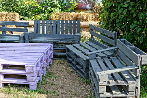 gray benches and a purple table made of wooden pallets stand on the ground and green grass outdoors in a summer park