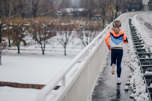 Rear view of an athletic woman wearing a headphones and listening music while jogging on a bridge with snow on cold winter day. Winter fitness. Fit sportswoman running in the city on snow at winter.