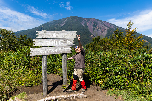 A senior Japanese woman stands by a mountain summit marker after her mountain hike.