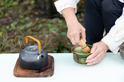 A close up of a senior Japanese woman making matcha tea the traditional way outdoors at a campsite.