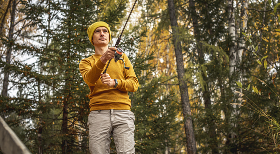 Young man fisherman in a yellow sweater and hat is fishing on the river bank in the autumn forest with a spinning rod in his hands. Concept of relaxing outdoor recreation, lifestyle, hobby