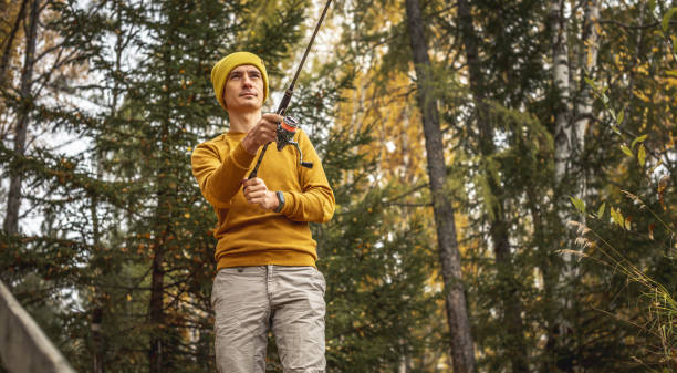 Man in a yellow sweater and hat is fishing on the river bank in the autumn forest with a spinning rod in his hands stock photo