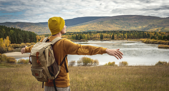 Tourist in a yellow hat and sweater with a backpack is standing with his arms outstretched to the sides in front of a river, the autumn golden forest and the hills. Concept of freedom, travel, hiking
