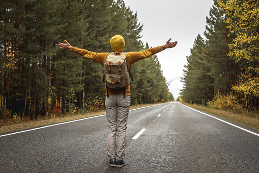 Man in a yellow hat and a sweater on a smooth empty asphalt road along a beautiful golden forest is standing with his arms outstretched to the sides. Concept of freedom, autumn, travel, hiking