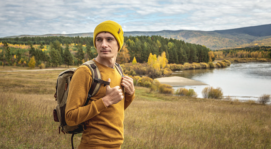 Man traveler in a yellow hat and sweater with a backpack is standing in front of the river, the autumn golden forest and the hills. Concept of freedom, travel, hiking