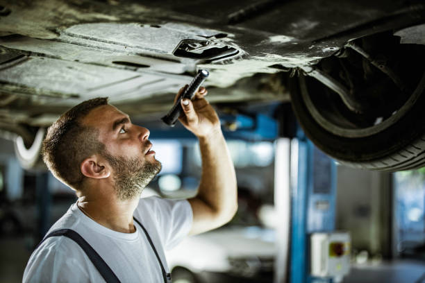 junger automechaniker, der das fahrwerk eines autos in einer werkstatt untersucht. - maintenance engineer car lamp protective workwear stock-fotos und bilder